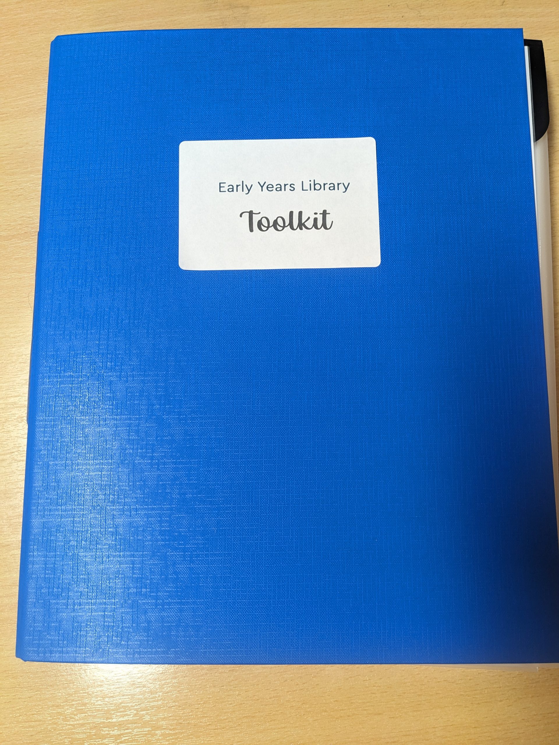 Blue binder on a wooden desk with a white label that says 'Early Years Library Toolkit'.